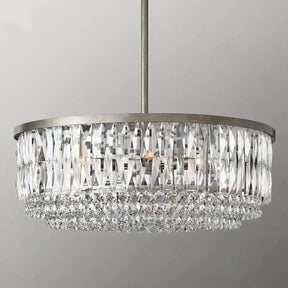 The Bacci Crystal Chandelier by Morsale.com is a modern cylindrical luxury chandelier, featuring multiple tiers of hanging K9 crystal prisms and larger droplets at the bottom. It is suspended from the ceiling by a metallic rod and lit internally, casting a sparkling, elegant glow.