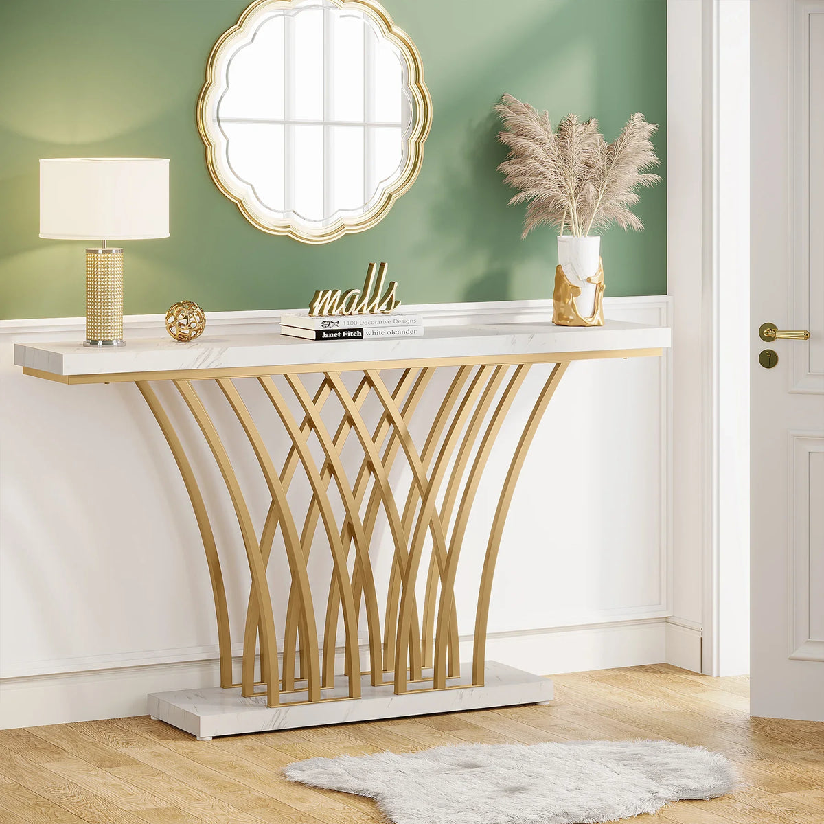 The elegant entryway features a modern Morsale Bassett Entryway Table with an arched gold base, adorned with a lamp, pampas grass, and ornaments. A round gold mirror is mounted on the green wall above. Light wooden floors and a small rug complement the space alongside a visible white door.