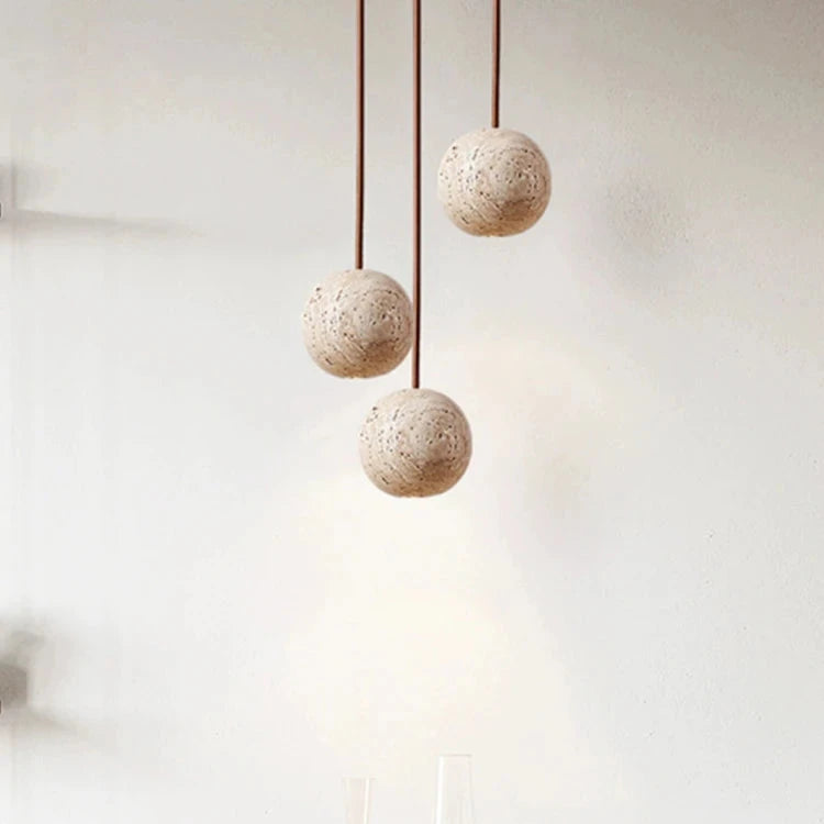 The Travertine Ball Pendant Light Fixture from Morsale.com, crafted from natural travertine stone, features a beige, porous surface that exudes a rustic and organic charm as it hangs from the ceiling. This elegant light fixture is suspended by a brown cord against a plain, off-white background.