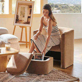 A woman in a striped dress lifts the lid of a Morsale Barrel Accent Chair's matching ottoman, revealing its chemical-free storage. She sits on the brown armchair, amid modern decor like a wooden table, woven rug, with an outdoor view and an easel in the background.