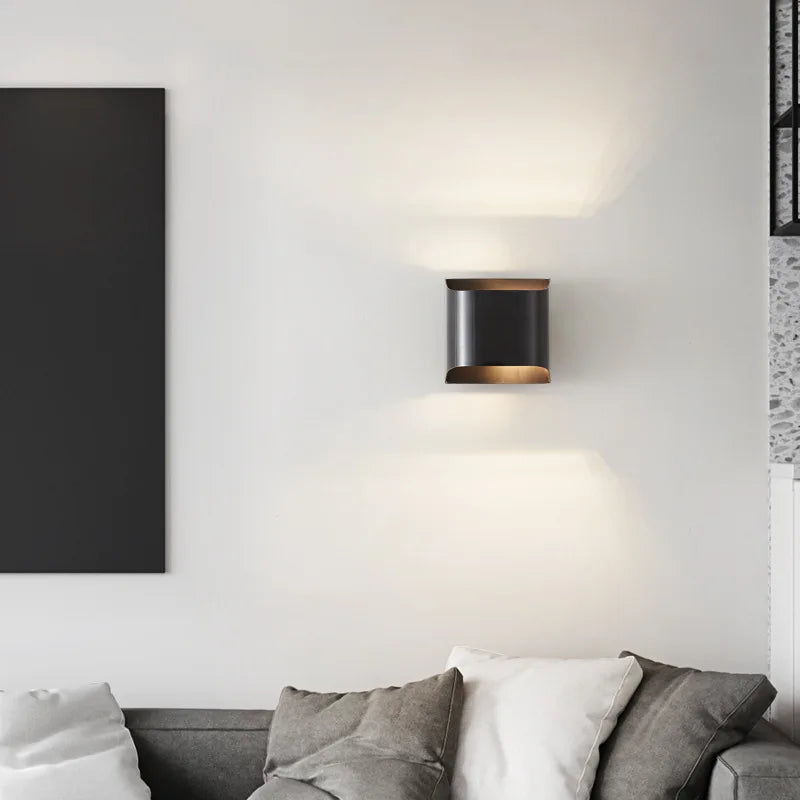 A modern living room showcasing a gray sofa adorned with white and gray cushions, where the Morsale Rame Copper Sconce elegantly illuminates the light gray wall above and below. The sleek LED wall lamp complements the partially visible large black rectangular artwork, while subtle brass accents enhance the overall elegance of the space.