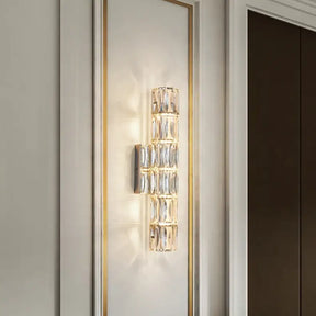 The Bacci Crystal Wall Light Sconce from Morsale.com features a modern vertical arrangement of handmade crystal blocks that emit a warm, ambient light. Mounted on a white wall with gold trim, this fixture adds an elegant touch to the minimalist living room décor.