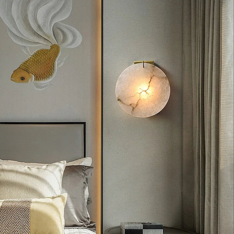 The bedroom corner features the Morsale Moonshade Marble Wall Lamp with LED lighting, casting a warm glow on the decorative fish art piece. The bed is adorned with white and beige pillows and a yellow one. Partially visible curtains on the right add to the modern style ambiance.