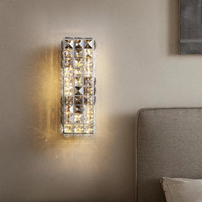 A Morsale Bacci Smoke & Clear Crystal Wall Sconce, with an integrated LED feature, emits a warm, glowing light, casting reflections on a beige wall. It is mounted above a wooden nightstand with a tray holding dark items. Part of a beige upholstered headboard and bed with white bedding is visible.