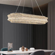 A modern dining room features a Bacci Modern Crystal Chandelier by Morsale with LED bulbs hanging over a marble table, adorned with decorations and surrounded by elegant chairs. Dark paneled walls and long curtains enhance the sophisticated ambiance.