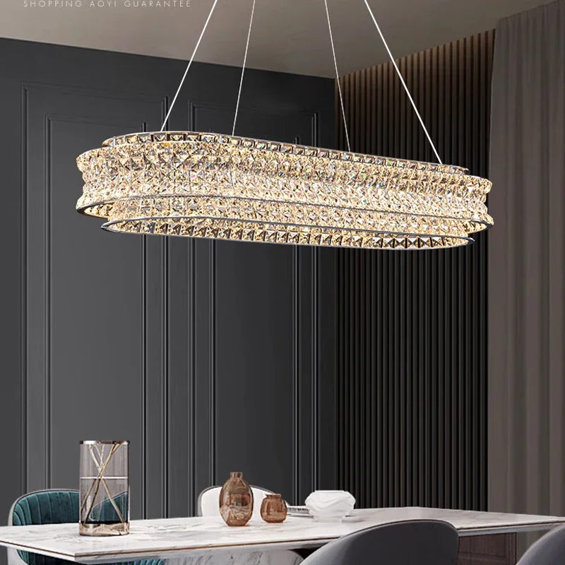 A modern dining room features a Bacci Modern Crystal Chandelier by Morsale with LED bulbs hanging over a marble table, adorned with decorations and surrounded by elegant chairs. Dark paneled walls and long curtains enhance the sophisticated ambiance.