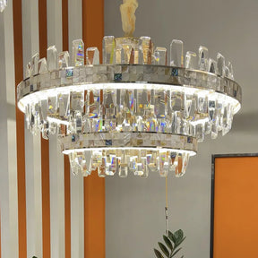 The luxurious Calvi Crystal Chandelier by Morsale is a modern light fixture featuring two circular tiers embellished with crystal prisms that cast a bright, warm glow. Complemented by orange and gray walls with vertical white stripes and surrounded by green plants, it perfectly captures the essence of elegant modern style lighting.