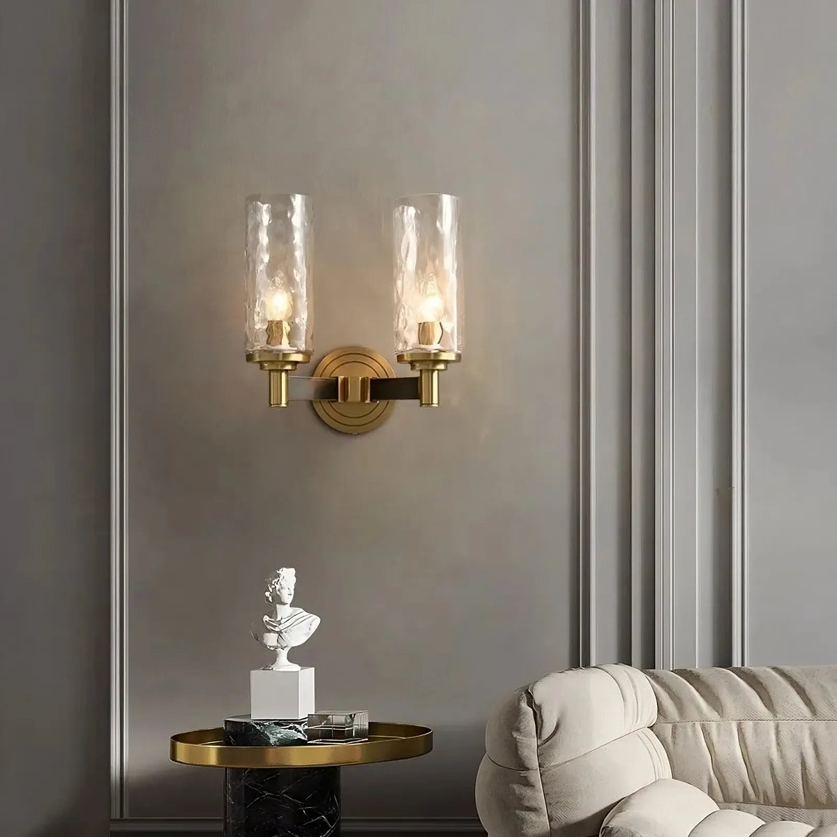 A modern interior featuring the Morsale Romea Copper Sconce, a wall-mounted dual light fixture with clear textured glass shades and LED bulbs that cast warm light. Below, a small marble table displays a copper bust sculpture next to a plush, light-colored sofa against a paneled wall.