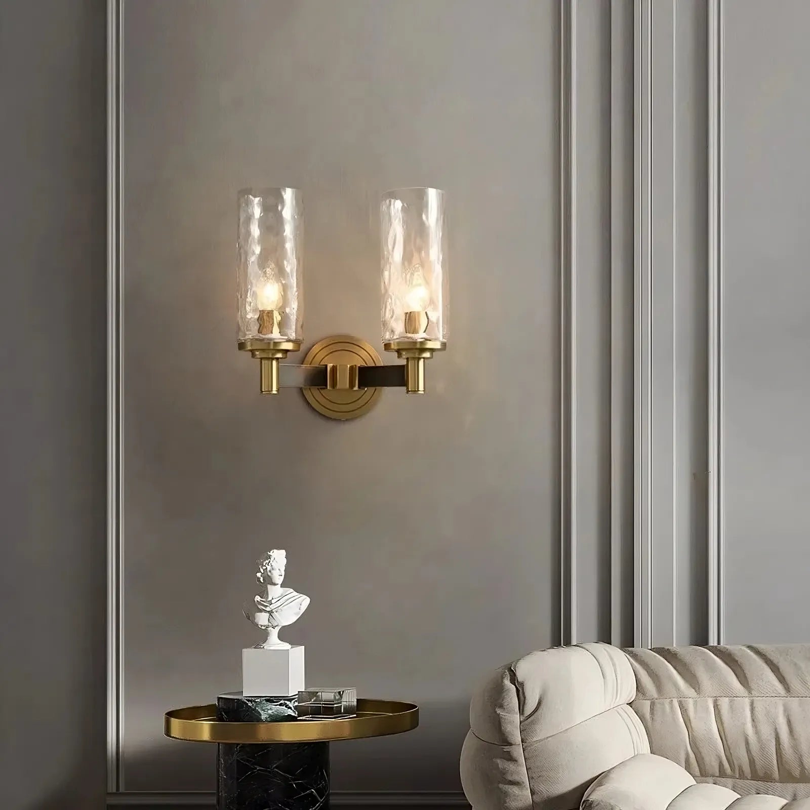 A modern interior featuring the Morsale Romea Copper Sconce, a wall-mounted dual light fixture with clear textured glass shades and LED bulbs that cast warm light. Below, a small marble table displays a copper bust sculpture next to a plush, light-colored sofa against a paneled wall.