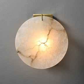 The Moonshade Marble Wall Lamp by Morsale, with a round marble texture and intricate veining, emits a soft warm glow. It features a modern style on a small gold bracket and uses LED for efficient illumination against a gray background.