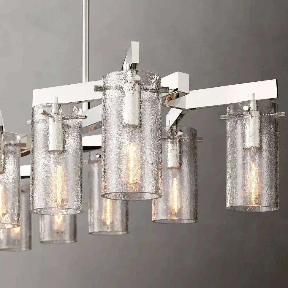 The Morsale Leroi Chrome Dining Room Chandelier features an elegant design with a metallic frame and cylindrical glass shades. Each shade houses an elongated light bulb, casting a warm glow through the textured glass. Versatile lighting hangs beautifully against a neutral gray background.