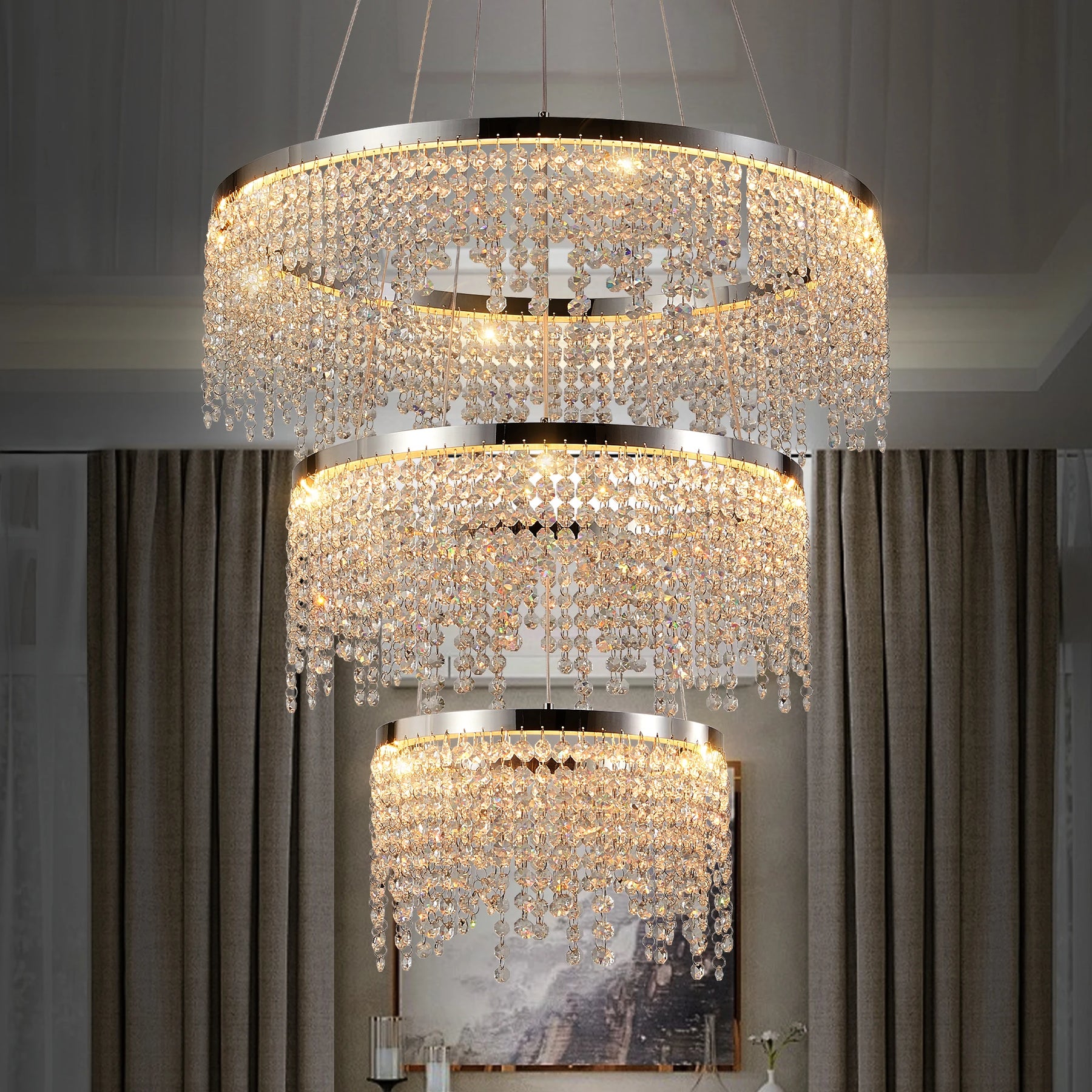 The Morsale Alleri 3-Tier Crystal Chandelier, featuring circular tiers of crystal beads and LED bulbs, emits a warm, elegant glow. It complements modern decor elements like curtains and framed paintings beautifully in any stylishly curated space.