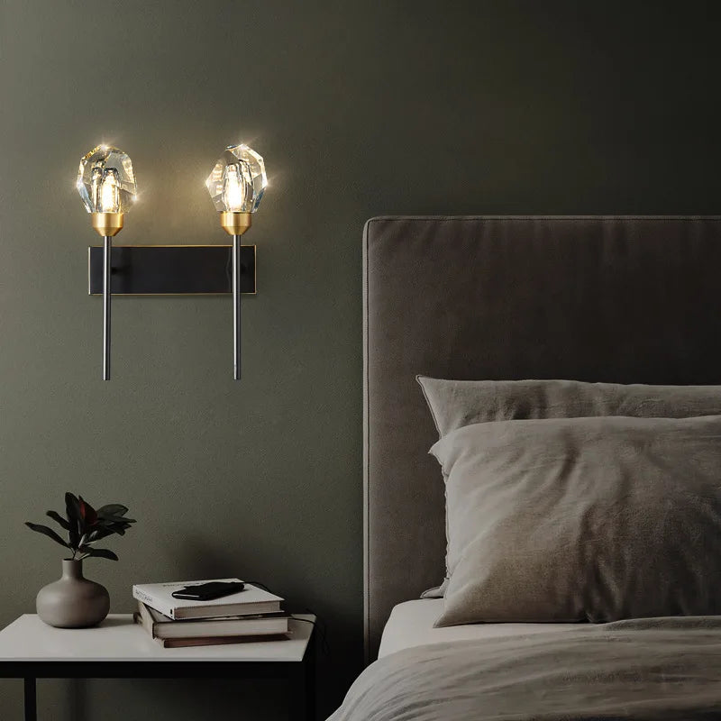 A modern bedroom features the Morsale Diamante Crystal Wall Sconce, boasting two illuminated glass bulbs and copper accents. The bed, dressed in neutral-toned bedding, is positioned next to a small table decorated with a plant and books, all set against a dark, minimalist wall.