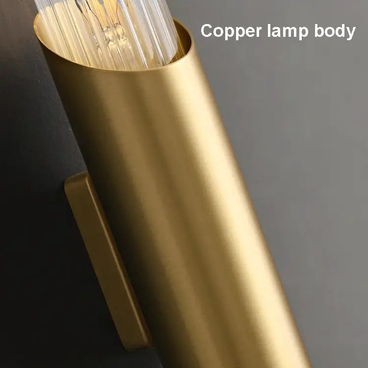 Close-up image of an elegant lighting fixture with a lit cylindrical copper wall sconce and a visible filament bulb inside. The caption "Palermo 23\" Brass Wall Sconce Light by Morsale" is written in white text on the upper right corner of the image. The modern design lamp is mounted on a smooth, gray wall.