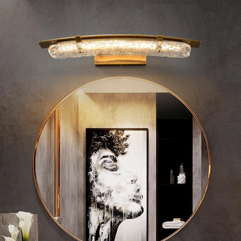 A modern Copper & Glass LED Wall Sconce by Morsale is mounted above a round mirror. Its textured glass shade and contemporary design illuminate an artistic black-and-white painting of a woman's face, enhancing the stylish and contemporary ambiance.