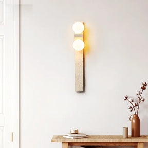 A modern Japan Style wall-mounted light fixture from Morsale.com, featuring two glowing circular LED bulbs, is vertically installed on a light-colored wall. Below the Natural Travertine Modern Wall Lights, to the left, a green leafy plant is partially visible.