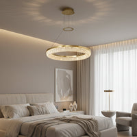 A modern bedroom showcases the Bergamo Modern Chandelier by Morsale.com, a gold circular LED fixture above a neatly made bed. Large windows with sheer curtains brighten the space, while neutral walls add to the contemporary ambiance.