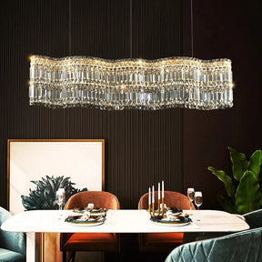 A dining room with a white table set for a meal, featuring wine glasses and a candle holder with white candles. Above the table hangs the elegant, elongated Bacci Crystal Chandelier For Dining Room by Morsale.com. The room has dark, textured walls, and there are two brown chairs and two teal chairs around the table. A framed painting and a large green plant are in the background.