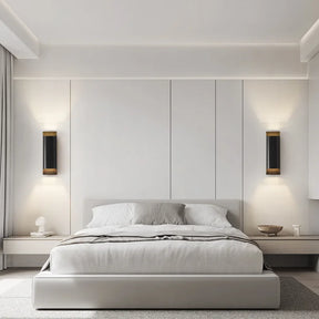 A contemporary bedroom showcasing a minimalist aesthetic includes a low-profile bed with white bedding and a sleek headboard. On either side, the room is illuminated by the Rame Copper Sconce from Morsale, paired with small bedside tables in neutral tones accented by subtle brass details.