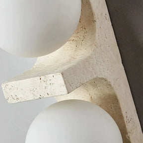 A modern Japan Style wall-mounted light fixture from Morsale.com, featuring two glowing circular LED bulbs, is vertically installed on a light-colored wall. Below the Natural Travertine Modern Wall Lights, to the left, a green leafy plant is partially visible.