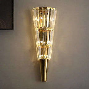 The Gio Stainless Steel & Crystal Sconce by Morsale is a contemporary LED wall fixture with a gold finish and crystal accents, creating a warm glow. It features a tapered design, beautifully mounted on stainless steel against a neutral-toned wall.