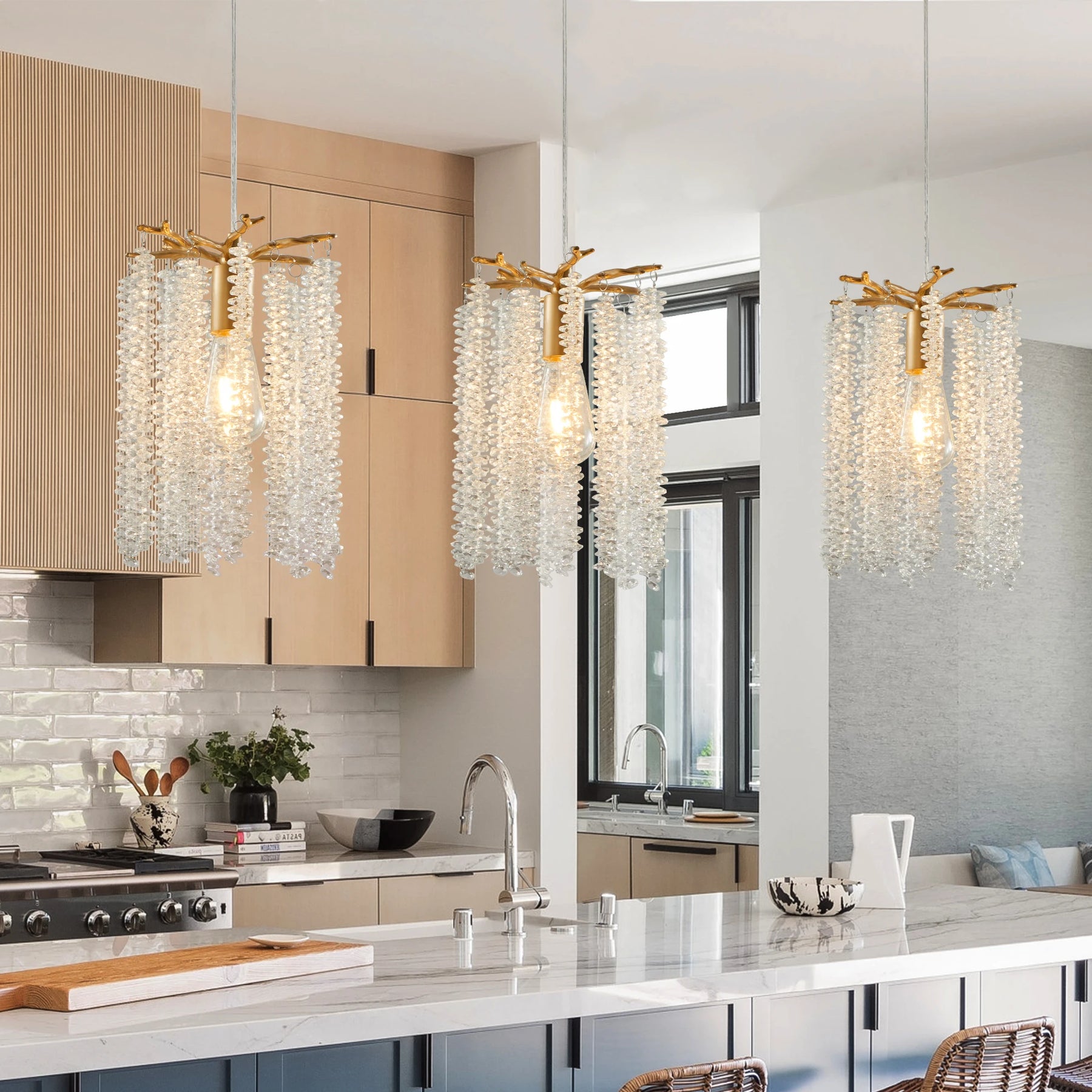 A modern kitchen showcases light wood cabinets, a marble island, and sleek stainless steel appliances. Above the island hang three Albero Crystal Pendant Lights by Morsale. Large windows flood this chic metal-accented space with natural light.