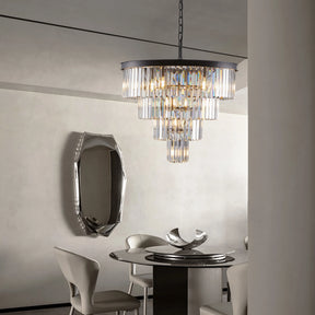 A modern dining room with a round table and four chairs exudes luxury. The 23.6" Gio Crystal Chandelier by Morsale sparkles elegantly from the ceiling, complemented by a decorative metallic bowl on the table and an abstract-shaped mirror on the wall.