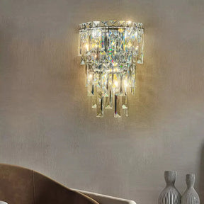 A Bacci Crystal Contemporary Wall Sconce by Morsale, featuring cascading crystal prisms, casts a warm, ambient light in a cozy living room. Below it, part of a curved beige sofa and two decorative gray vases are visible against a neutral-toned wall.