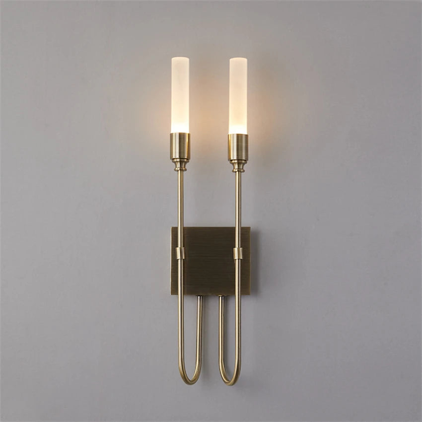 The Candela Contemporary Wall Sconce by Morsale is a modern lighting fixture featuring two sleek, cylindrical glass shades that emit a warm glow. With its minimalist design, metallic finish, and clean lines, it enhances interior design by providing elegant lighting against a plain gray background.