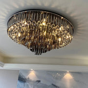 A luxurious, round Gio Crystal Ceiling Light by Morsale.com features cascading smoke grey crystals and multiple small lights, all mounted on a stainless steel ceiling fixture. The chandelier emits a warm, shimmering glow. Below it, a wall showcases a modern, abstract painting with shades of gray, white, and blue.