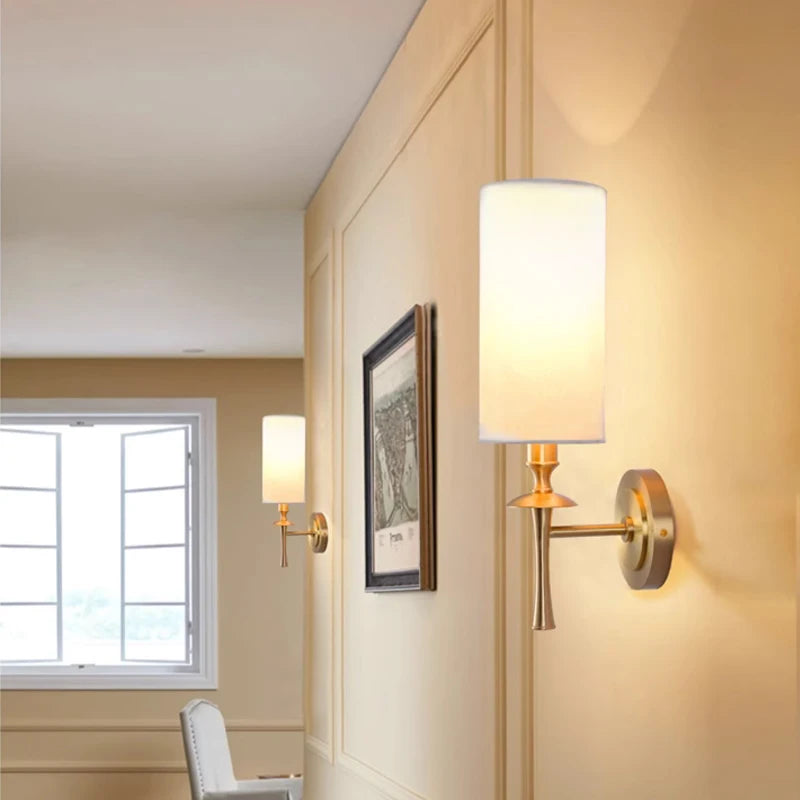 A warmly lit hallway showcases the Morsale American Retro Copper Wall Lights, featuring cylindrical white shades and elegant copper accents. An open window allows natural light to enhance the minimalist decor, while framed artwork on the beige wall is perfectly complemented by energy-efficient LED lighting.