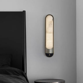 A modern bedroom showcases Morsale's Sandford Stainless Steel & Marble Wall Sconce above a black side table and sleek black headboard on a gray wall. The oval-shaped LED emits a warm glow, contrasting with the minimalist decor.