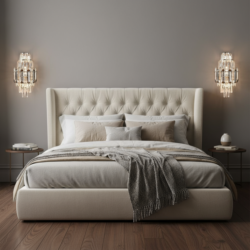 A modern bedroom showcases a tufted bed, neutral bedding, a gray throw, and round wooden nightstands. Morsale’s Lazzo Crystal Wall Sconces bring elegance to the gray wall above warm wood floors.