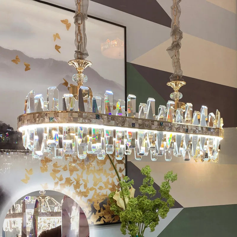 The Calvi Crystal Chandelier by Morsale, a quintessential luxury light fixture, hangs from the ceiling with intricate prismatic details. This modern style lighting is complemented by mirrored walls and subtle decorative elements, enhanced by some greenery visible below.