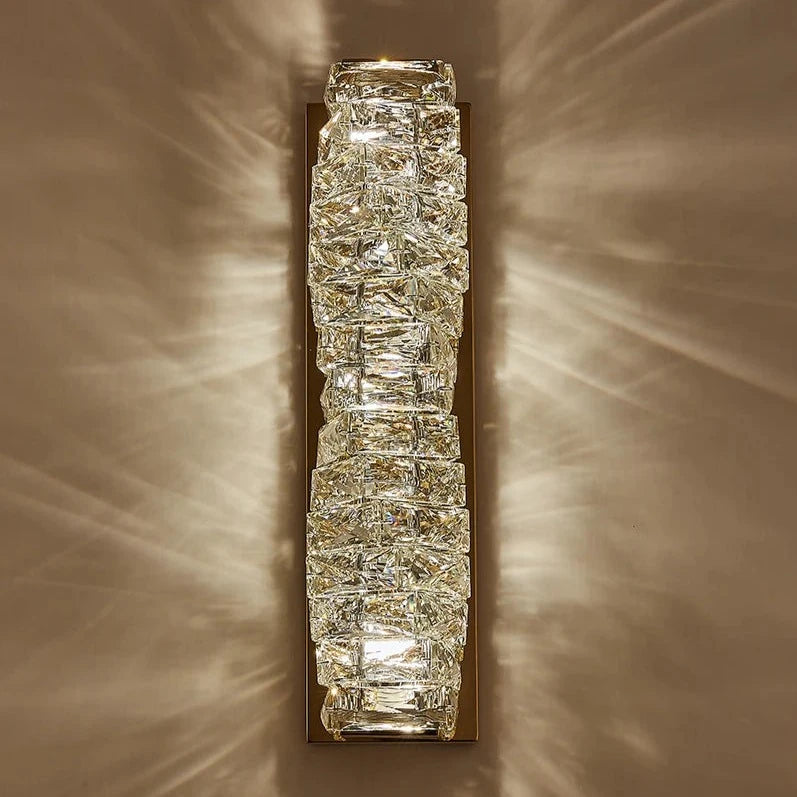 A vertical wall-mounted Morsale.com Bacci wall sconce with a gold frame and multiple rectangular, textured glass pieces emitting a warm, radiant light, casting a starburst pattern on the wall.