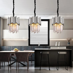 A modern kitchen features sleek cabinetry, a central island with two black bar stools, and a dining table with brown chairs. Above the island hang three Gio Crystal Kitchen Light Fixtures from Morsale.com, adorned with K9 Crystal pieces, adding a touch of sophistication and elegance to the stainless steel-accented space.