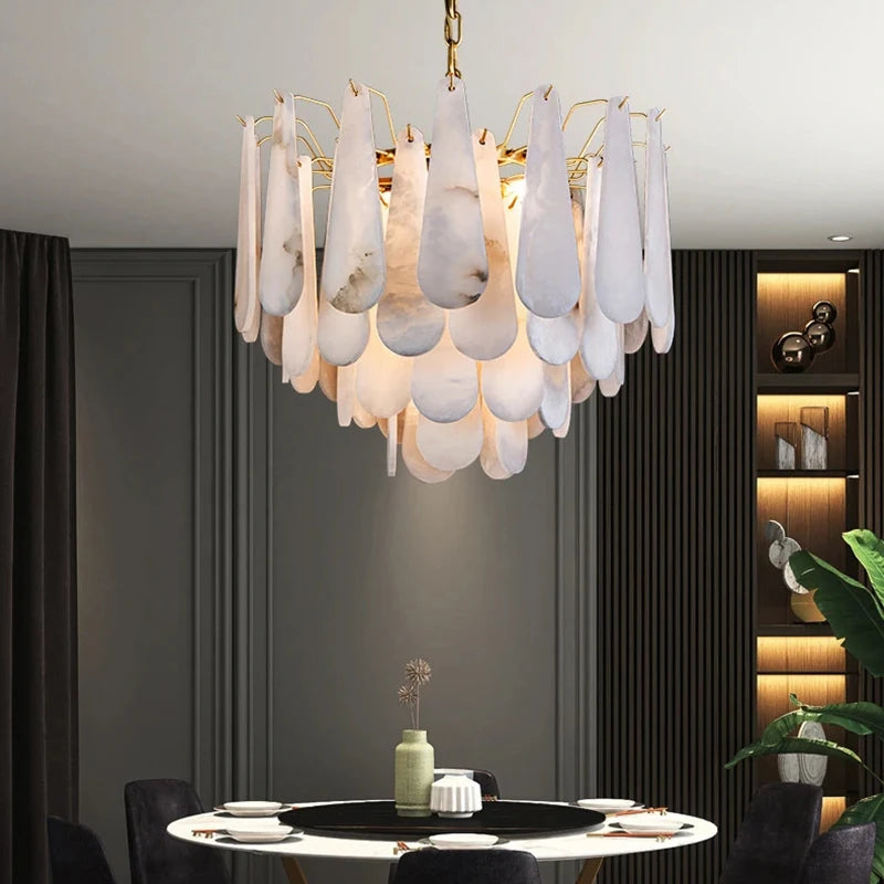 A contemporary dining room is highlighted by the Morsale Moonshade Marble Chandelier, which boasts cascading white, petal-like elements. Below, a round table is adorned with a vase and dishes. In the background, shelves hold decorative items, complemented by a large green plant that enhances the natural ambiance.