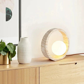 The Modern Travertine LED Table Lamp by Morsale.com features a round Natural Yellow Cave Stone frame with a textured, natural finish and a glowing spherical light bulb in the center. The lamp emits a warm, ambient light, creating a cozy atmosphere, and has a cord extending from the back.