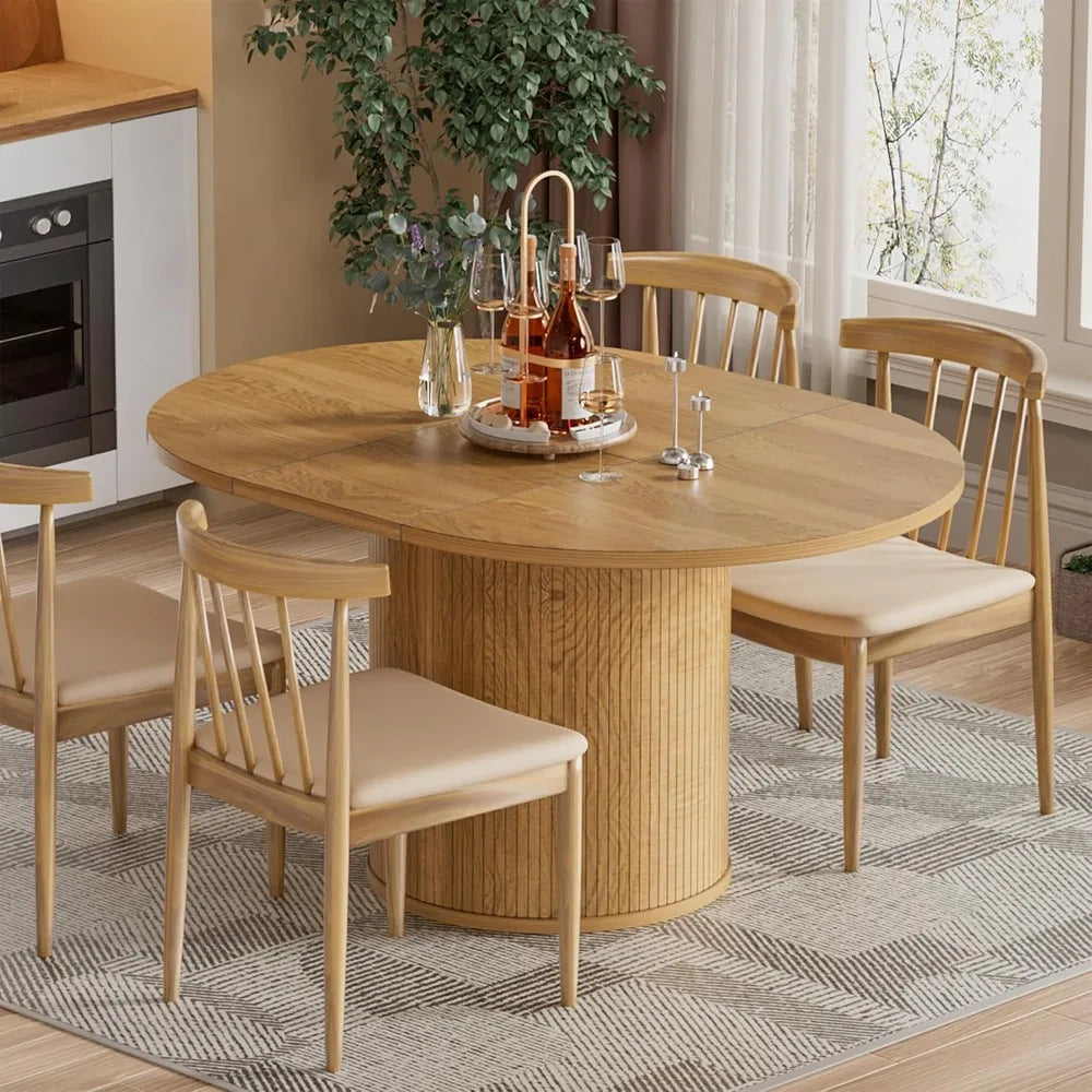 A modern dining area showcasing the Morsale Barrow Extendable Dining Table, paired with four beige cushioned chairs and a centerpiece of glasses with a carafe. The stylish setting is complemented by a kitchen backdrop, large window, and leafy plant for added coziness.