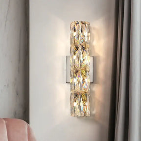 The Bacci Crystal Wall Light Sconce from Morsale.com, featuring modern LED technology and rectangular, handmade crystals, is mounted on a light gray wall. The sconce radiates a warm glow, producing reflections and patterns on the wall. Part of a blush pink upholstered chair and a gray curtain are also visible.