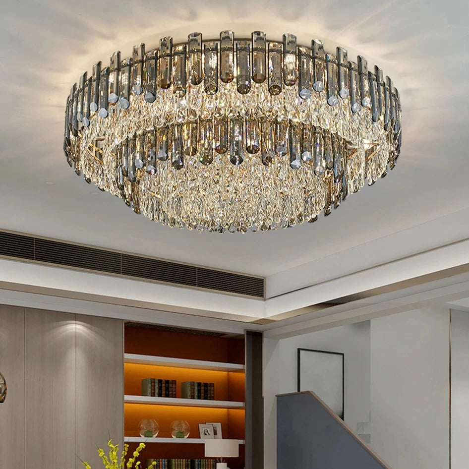 A modern, luxurious living room featuring a large handmade Venice 2-Tier Crystal Chandelier from Morsale.com adorned with crystal-like pendants. The room has a beige color palette with a comfortable sofa, a yellow floral centerpiece, shelves with decorative items, and an adjacent staircase leading upstairs.