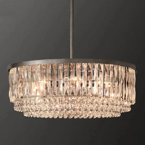 A round, luxurious Bacci Crystal Chandelier from Morsale.com featuring multiple tiers of hanging K9 crystals and beads, illuminated warmly against a dark, plain background. The chandelier, with its opulent gold finish, is suspended from a slender black rod.