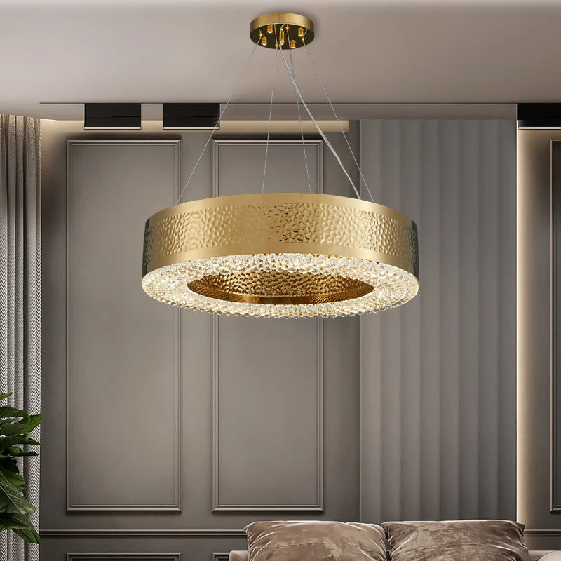 A modern living room showcases the Morsale Alleri Hammered Gold & Crystal Chandelier, featuring a textured surface and LED bulbs, elegantly suspended from the ceiling. The background includes a wall with subtle paneling next to a curtain, while part of a sofa is visible below.