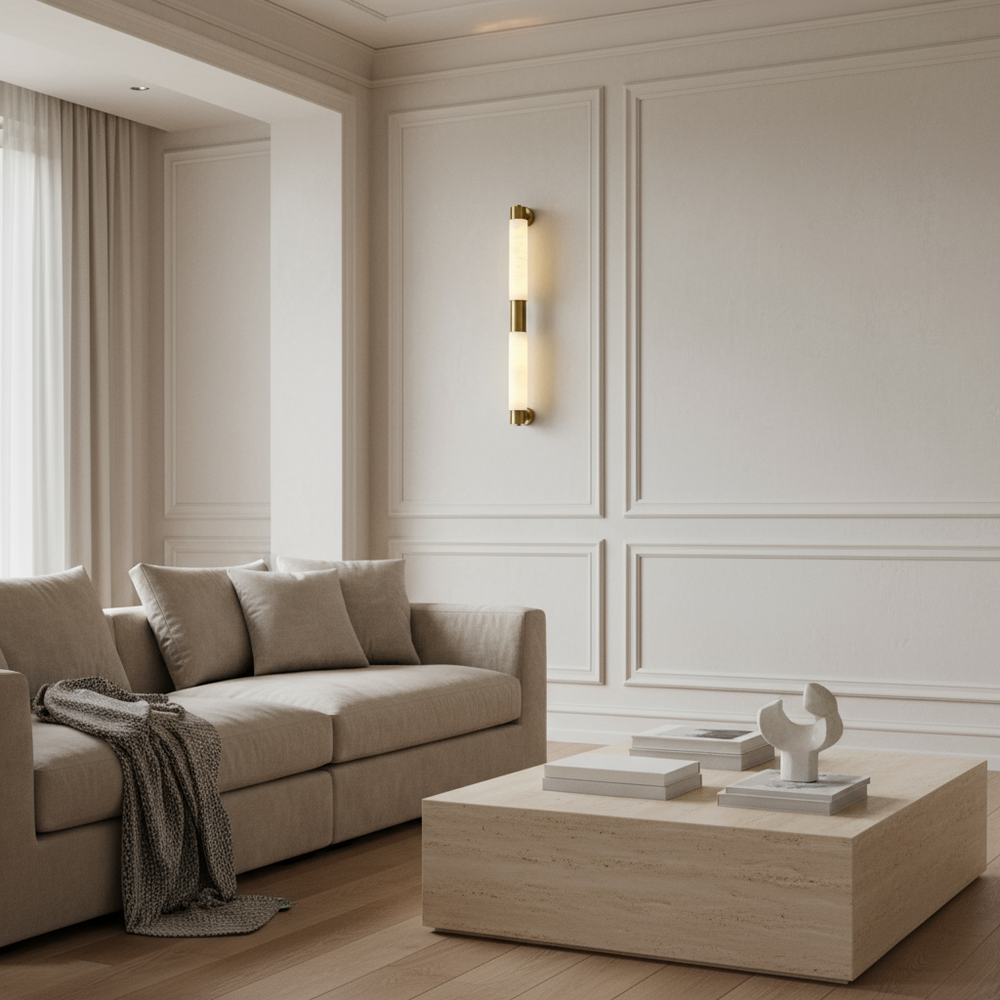 A minimal living room with a beige sofa, patterned throw, square stone coffee table with books and a modern sculpture, wood floors, cream-paneled walls, and the Maris Natural Marble Sconce by Morsale. The space is bright with natural light.