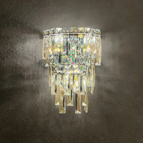 The Bacci Crystal Contemporary Wall Sconce by Morsale features a modern design with cascading prism-shaped crystals that emit vibrant reflections, beautifully complemented by an LED wall lamp and elegantly mounted on a textured dark wall.