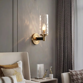 In a cozy bedroom corner, the Romea Copper Sconce from Morsale casts a warm glow above the beige upholstered headboard. The nightstand displays decorative items and a book, with LED bulbs creating soft shadows on the gray wall. Sheer curtains drape to the side, completing this serene space.