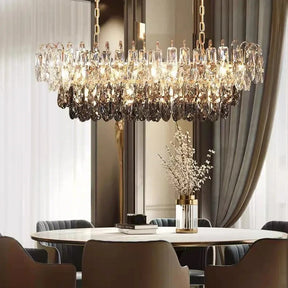 A luxurious dining room showcases a large oval table surrounded by sleek chairs, epitomizing modern style. Above the table hangs the Lazzo Contemporary Chandelier by Morsale, featuring LED bulbs that cast a gentle glow. At the center of the table is a vase with delicate white flowers, beautifully framed by soft natural light filtering through sheer curtains.