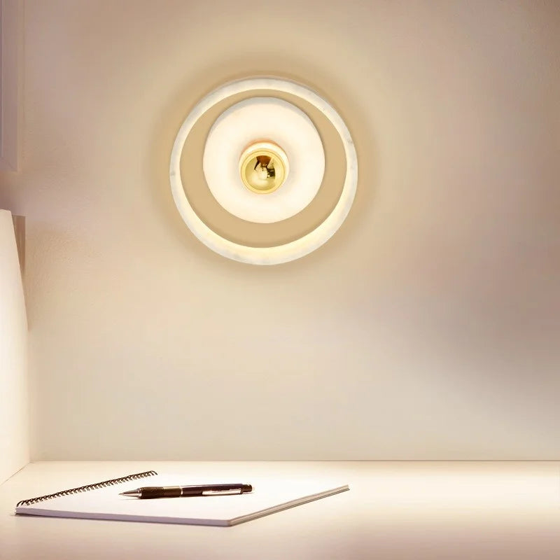 A Menards Modern Marble Wall Lamp by Morsale, featuring LED bulbs, emits a warm glow over a minimalistic desk. On the desk below, there is an open notebook with a black pen resting on it. The scene is simple and serene, with soft lighting creating a cozy atmosphere.