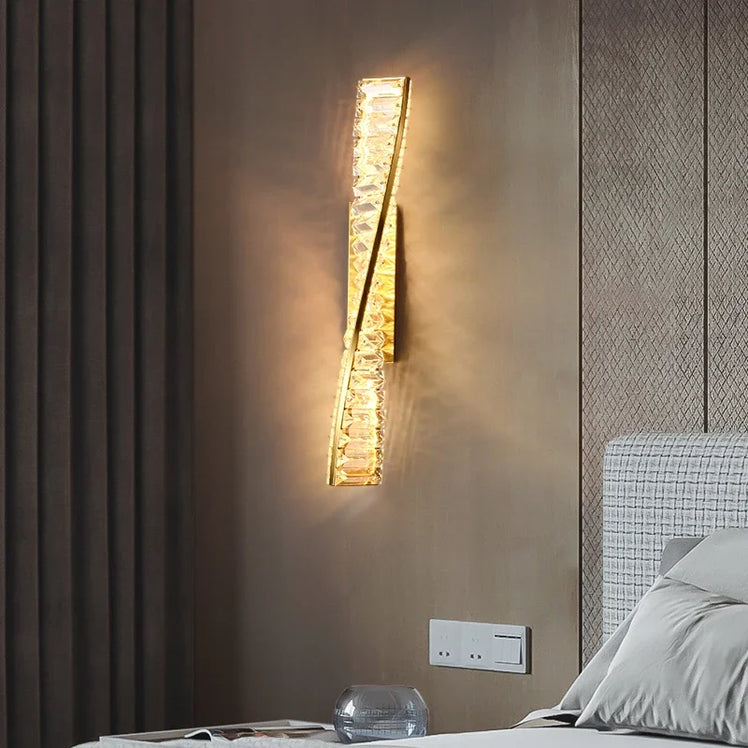 In a bedroom, the Bacci Crystal Wall Sconce by Morsale emits a warm, glowing light. This LED wall lamp showcases a vertically oriented, crystal-like design. The wall is adorned in neutral tones, with part of the bed featuring a light-colored headboard in view.
