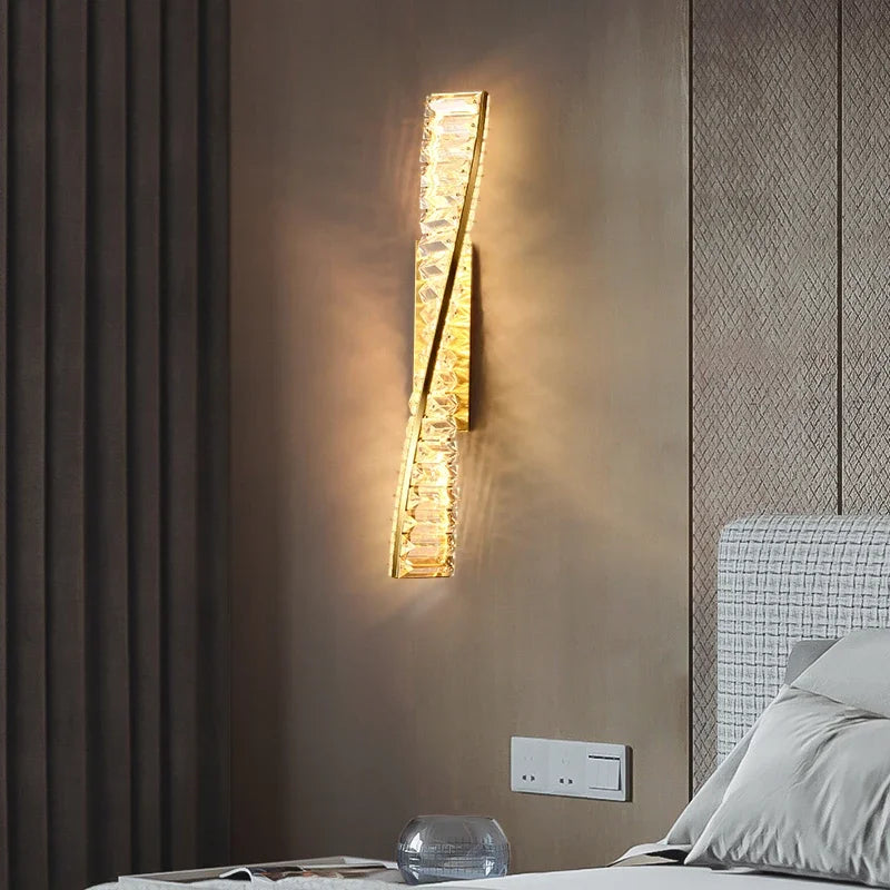 In a bedroom, the Bacci Crystal Wall Sconce by Morsale emits a warm, glowing light. This LED wall lamp showcases a vertically oriented, crystal-like design. The wall is adorned in neutral tones, with part of the bed featuring a light-colored headboard in view.